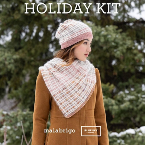 Person wearing a knitted hat and scarf with Christmas trees in the background, promoting a holiday kit by malabrigo.